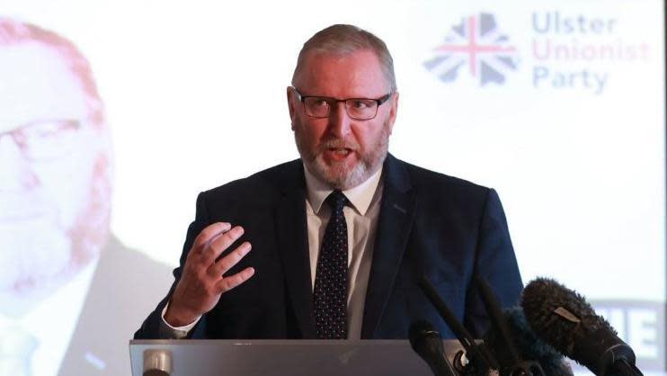 Doug Beattie standing at a podium and speaking at the Ulster Unionist Party manifesto launch. He is wearing a blue suit jacket, tie and white shirt.  