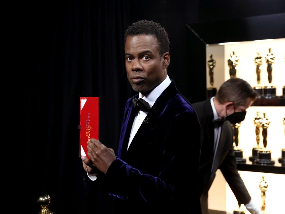 Chris Rock holding an Oscar winner envelope with Oscar statues behind him