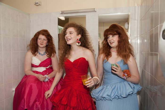 Three young ladies in prom dresses, each holding a drink, smiling in a bathroom. They have red and curly hair styled differently