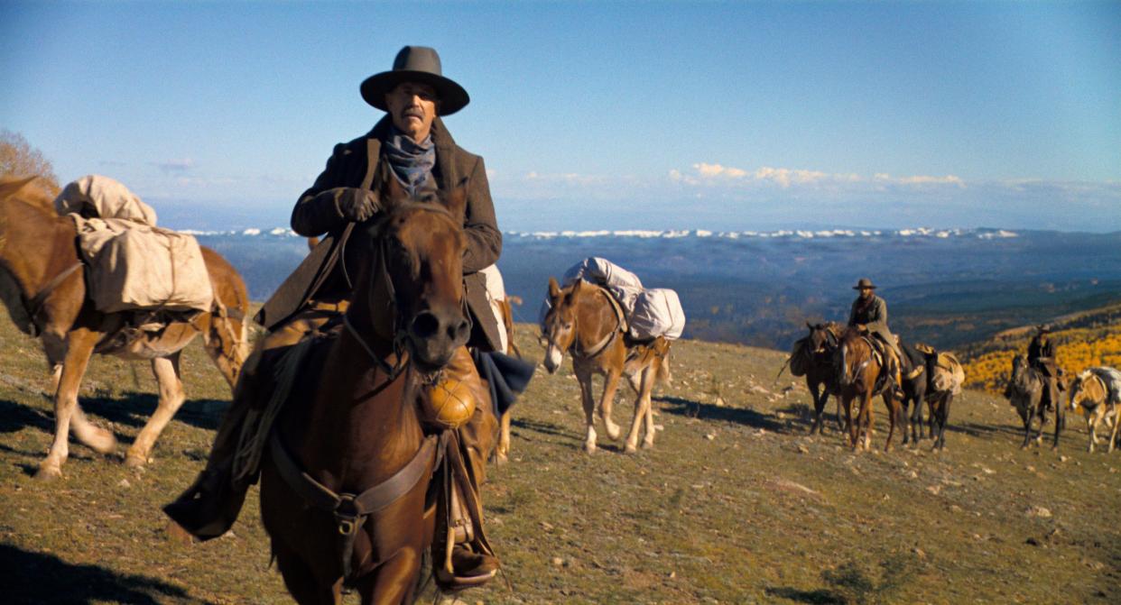 Kevin Costner, in cowboy attire, rides a horse with other horsemen around him.