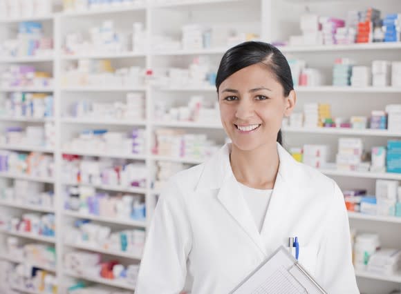 Woman in white lab coat holding clipboard in front of shelves of prescription drug bottles and containers.