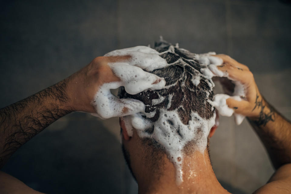 A person is seen from behind, washing their hair, with soap suds covering their head. The image focuses on the act of hair washing