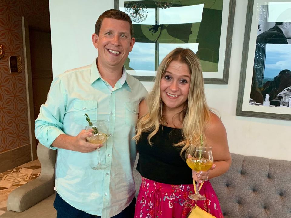 Photo of Terri Peters and her husband standing side-by-side with their arms around each other, holding cocktail glasses. Terri has long dyed blonde hair, brown eyes, and wears a black tank top with a bright pink, floral skirt. Her husband has short brown hair, hazel eyes and wears a green button down shirt. Behind them is a grey couch and framed art on the white walls.