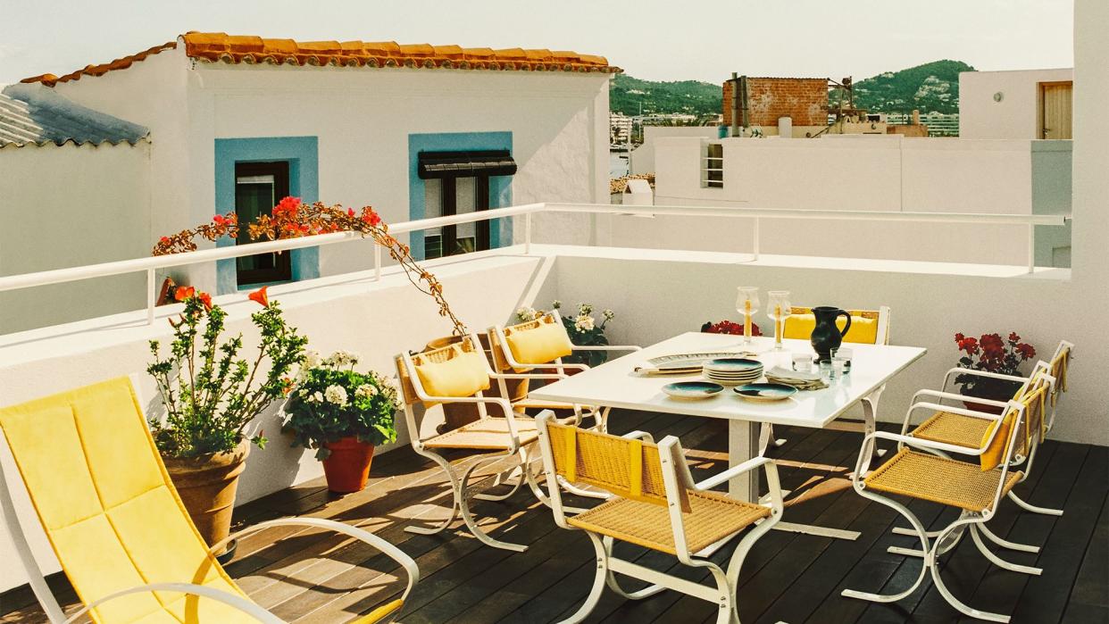 an open terrace with a dark stained deck with yellow back chairs and a white table set with dishes, potted flowering plants, a view of terracotta tiled roofs and mountains in distance