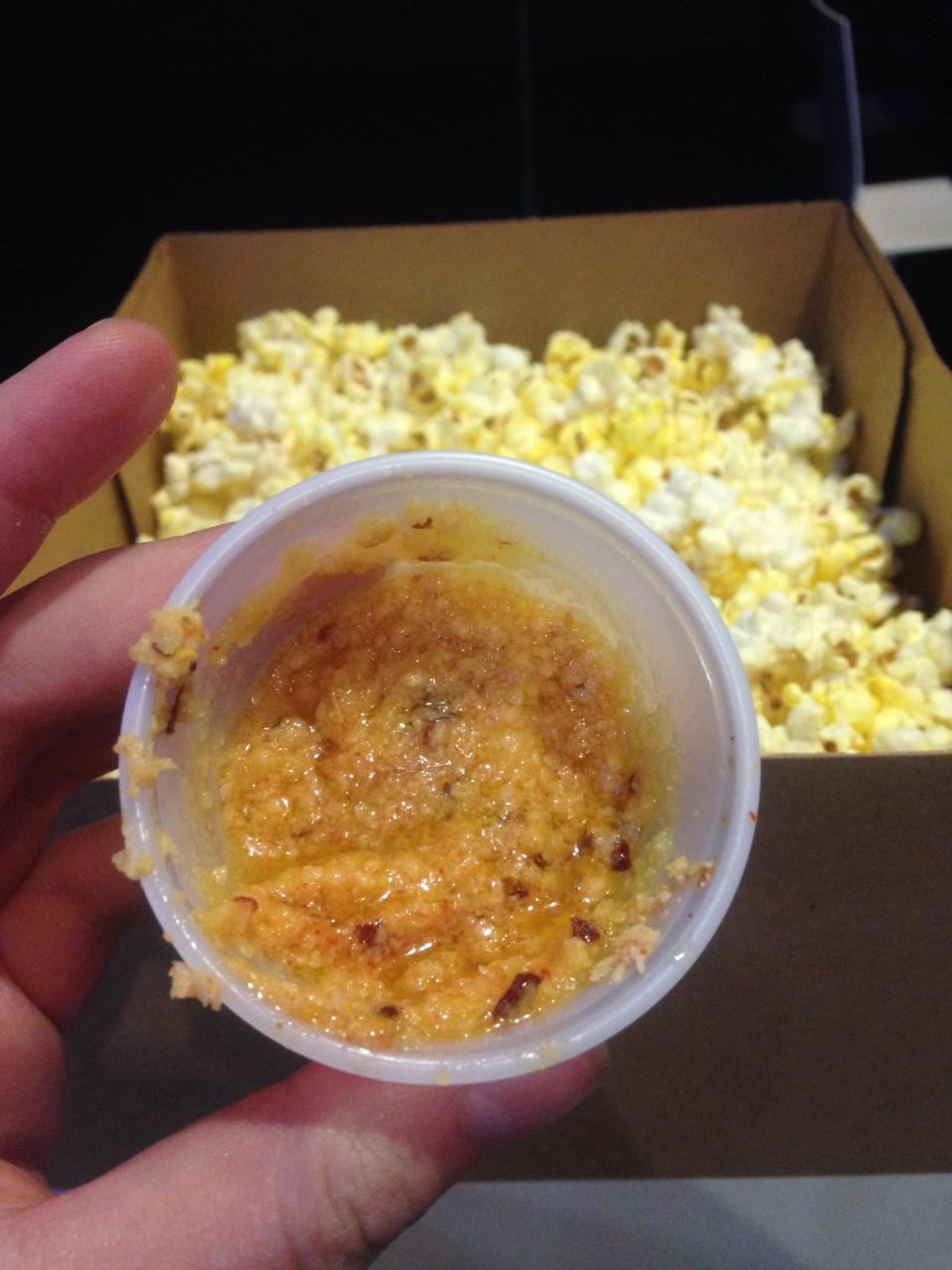 Hand holding a small cup of partially eaten orange-flavored ice beside a box of popcorn