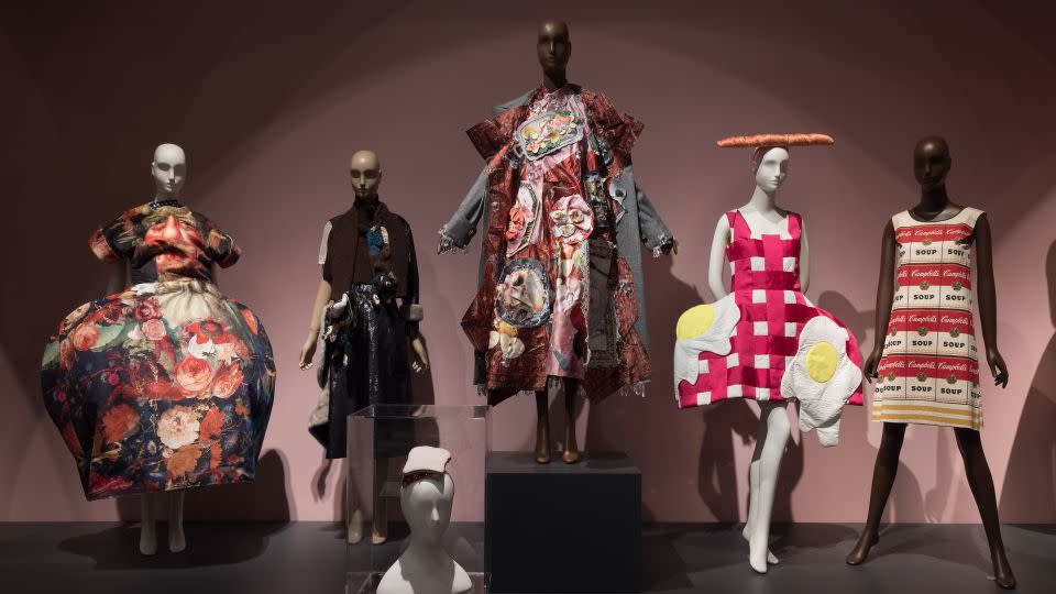Pieces included in the exhibition's "Feeding the Eye" display include a sculptural gown from Comme des Garçons Spring-Summer 2018 collection (far left),  a PB&J sandwich hat designed by Stephen Jones (foreground center) and the paper "Souper Dress" designed by Campbell's Soup Company circa-1966 (far right). - The Museum at FIT