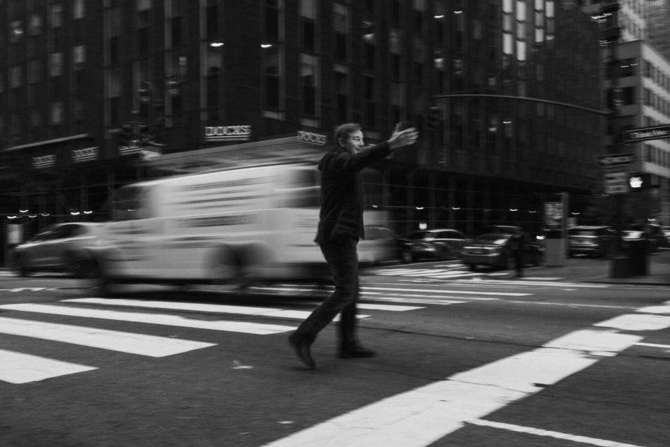 A black-and-white photo of a man standing in the crosswalk of a busy New York street as a car passes by.