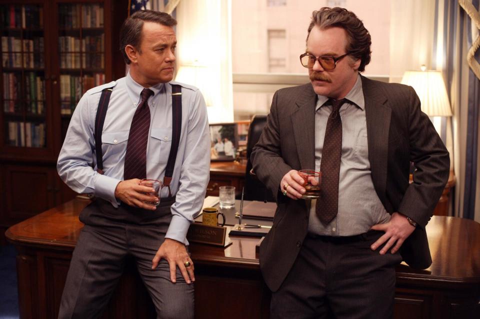 Two men in suits, one without his jacket, lean against a desk talking