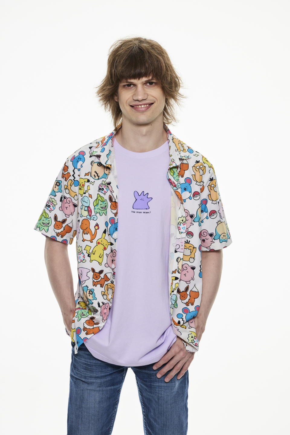 A man with brown hair stands smiling on a white background in blue jeans, lavender Ditto tee and a white shirt featuring a vast array of colourful Pokemon.