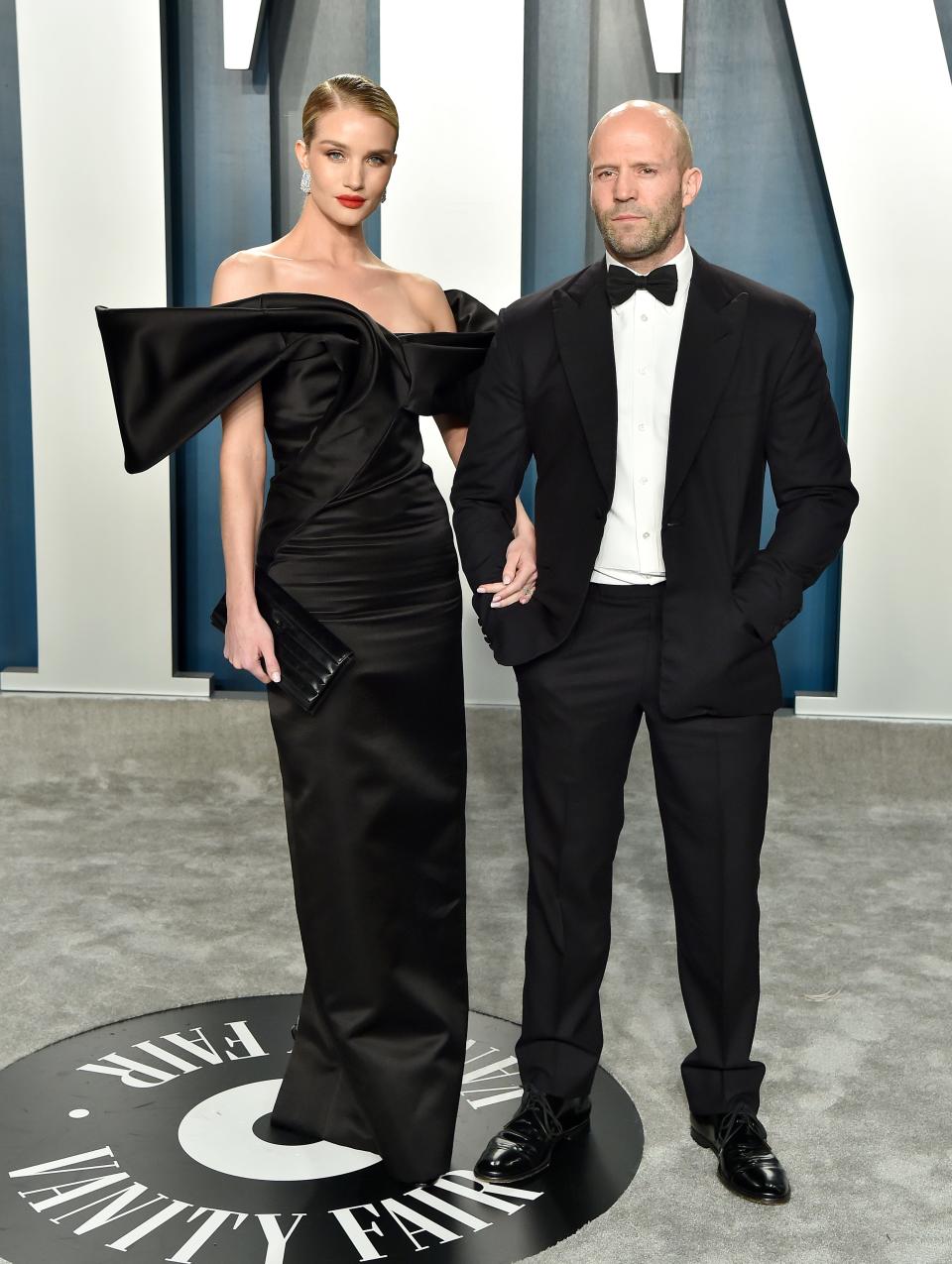 Rosie Huntington-Whiteley in a black floor-length gown standing taller than Jason Statham in a tuxedo.