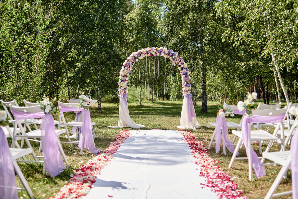 Outdoor wedding setup with an arch adorned with flowers and a petal-lined aisle surrounded by white chairs covered with fabric in a tranquil garden setting