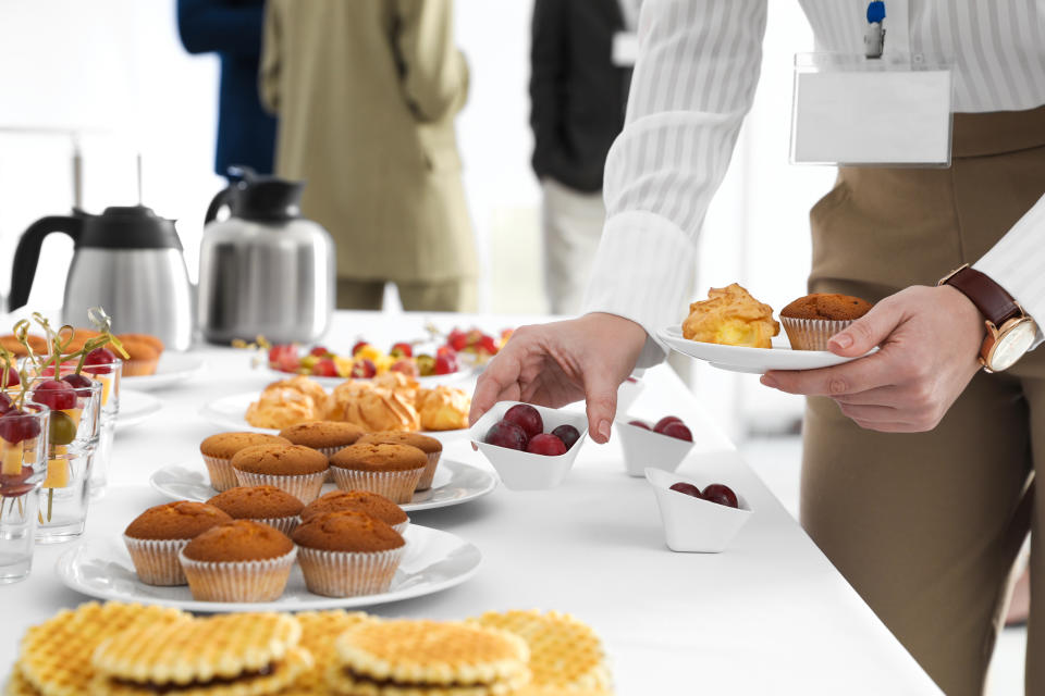 Person serving themselves from a buffet table with muffins, grapes, pastries, and sandwiches at what appears to be an event or meeting