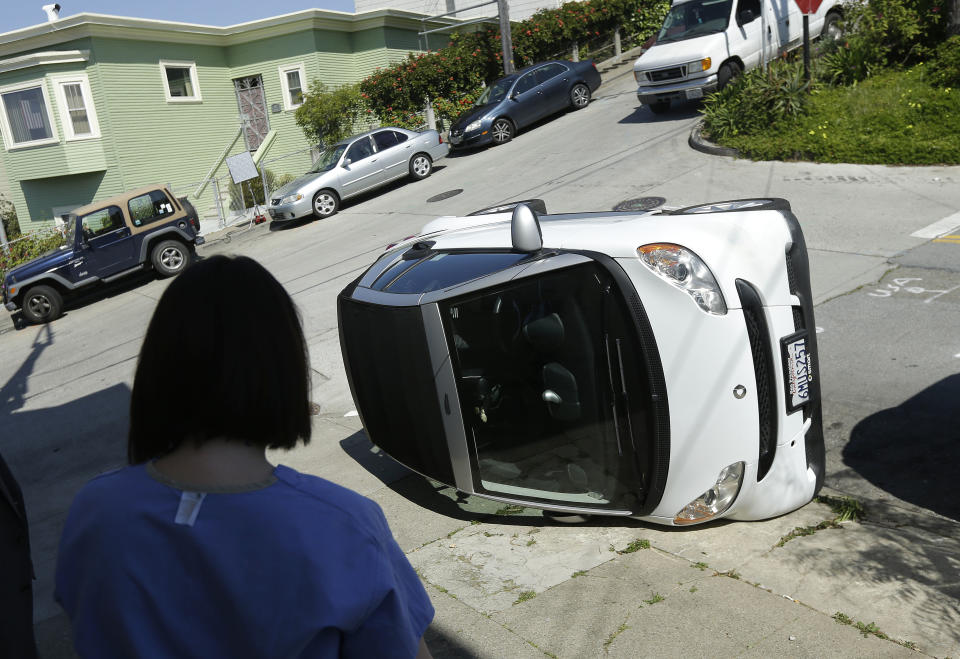 Shelley Gallivan looks toward a tipped over Smart car which belongs to her friend on the corner of Prospect and Oso streets in San Francisco, Monday, April 7, 2014. Police in San Francisco are investigating why four Smart cars were flipped over during an apparent early morning vandalism spree. Officer Gordon Shyy, a police spokesman, says the first car was found flipped on its roof and a second was spotted on its side around 1 a.m. Monday in the Bernal Heights neighborhood. (AP Photo/Jeff Chiu)