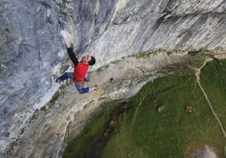 Tom Randall throws for a chalk stained hold while leading an overhanding grey cliff. The green valley and his belay look on from below.