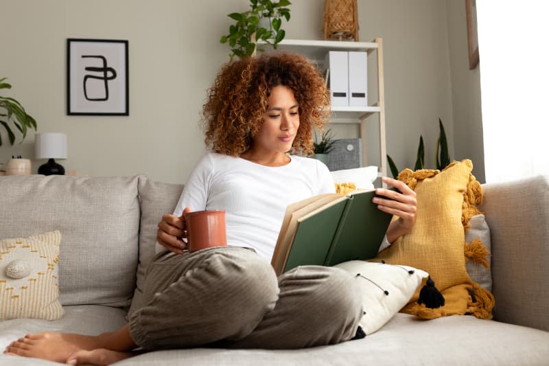Young multiracial woman reading a book at home, drinking tea sitting on the sofa in cozy living room. Lifestyle concept.