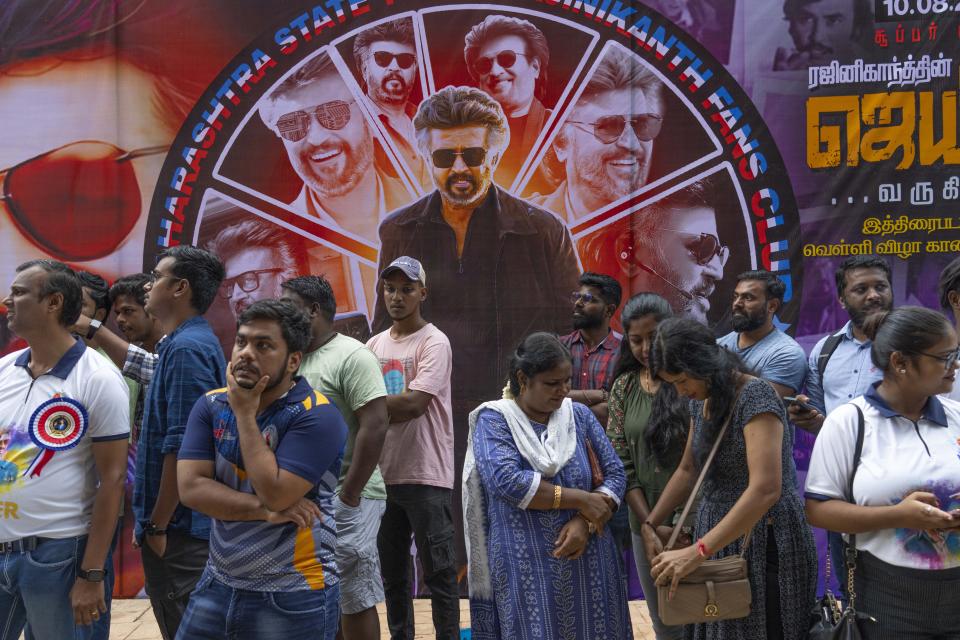 Fans wait near a poster of Indian superstar Rajinikanth, outside a cinema hall after they arrive to watch the screening of his latest film "Jailer" in Mumbai, India, Thursday, Aug. 10, 2023. (AP Photo/Rafiq Maqbool)