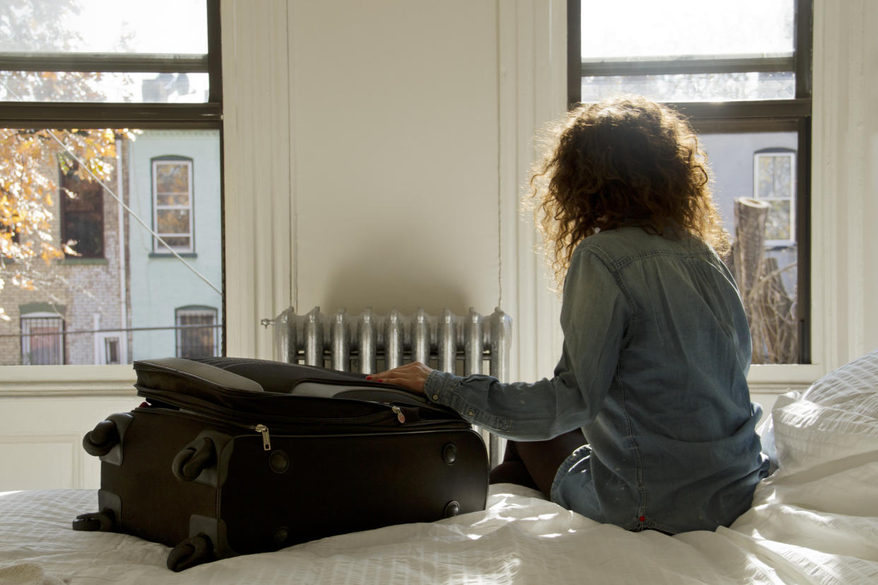 Female solo traveller sat on bed with suitcase looking out of the window in Brooklyn, New York.