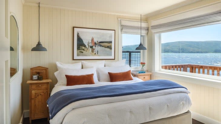 A queen-size bed covered with white covers and a sea-blue blanket, with a window view out to a sparkling bay and the green hill of Point Reyes in the distance