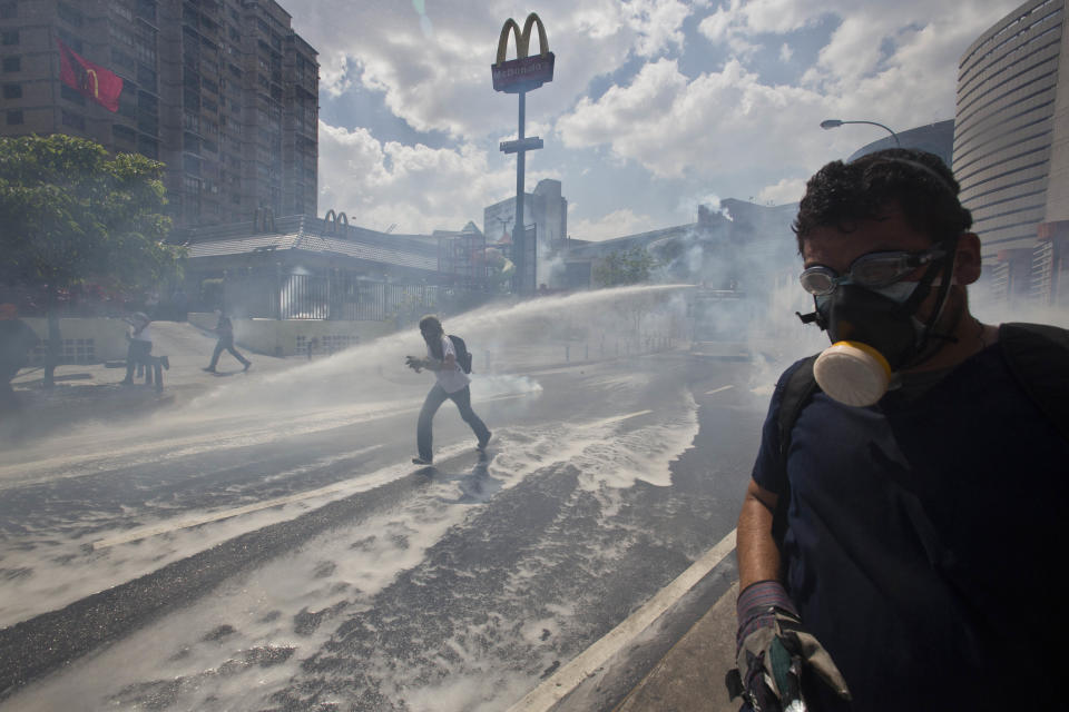 A masked protester runs from a spraying water canon operated by Bolivarian National Police in Caracas, Venezuela, Thursday, March 20, 2014. Thursday dawned with two more opposition politicians, San Cristobal Mayor Daniel Ceballos and San Diego Mayor Enzo Scarano, behind bars. Police used tear gas and water cannons to disperse a student-called protest of several thousand people in Caracas, some of those demonstrating against the arrests of the mayors. (AP Photo/Esteban Felix)