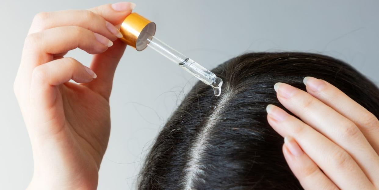 close up of a female head with dark hair woman using pipette with a cosmetic product near the hair parting the concept of dandruff