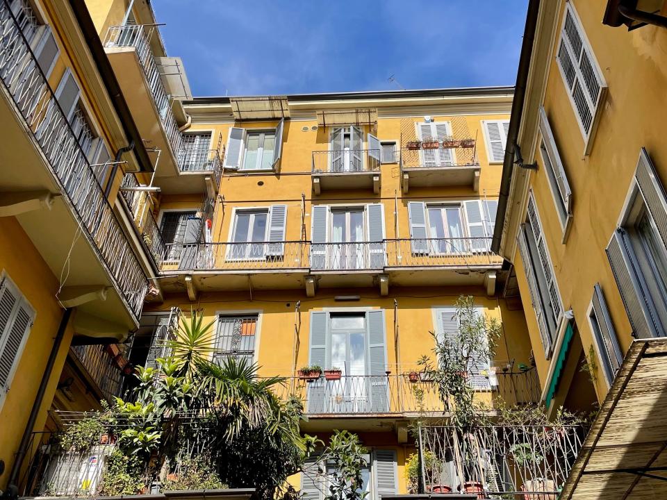 Photo from below of a yellow apartment building against a bright blue sky. The building has several plants on its lower balconies.