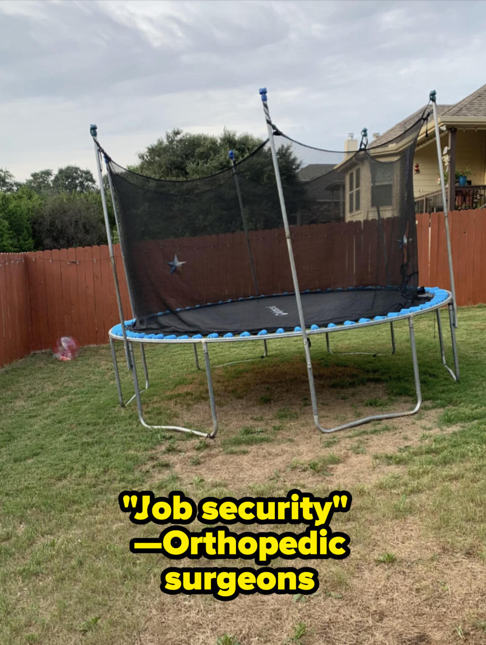 A trampoline with safety netting is set up in a backyard enclosed by a wooden fence, with a house in the background