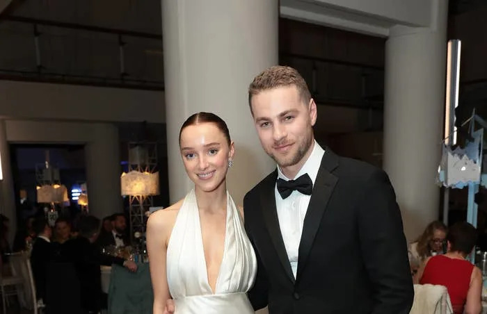 Phoebe Dynevor in a sleek, white halter-neck gown and Cameron Fuller in a black tuxedo, posing together at a formal event