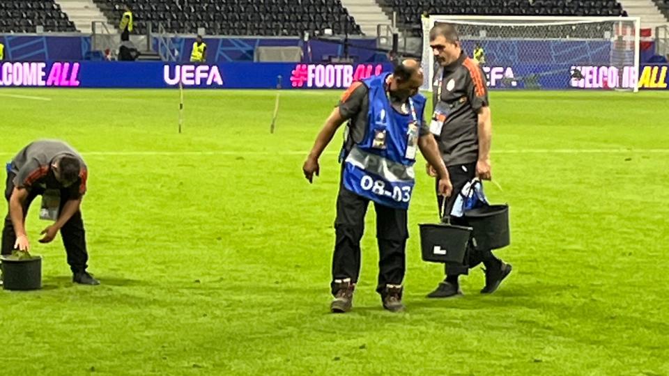 Three men hold buckets of grass on a football pitch