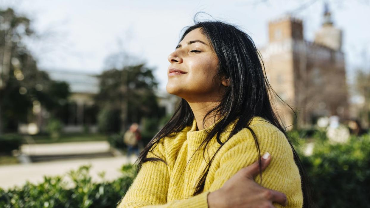 smiling woman hugging self against sky in public park