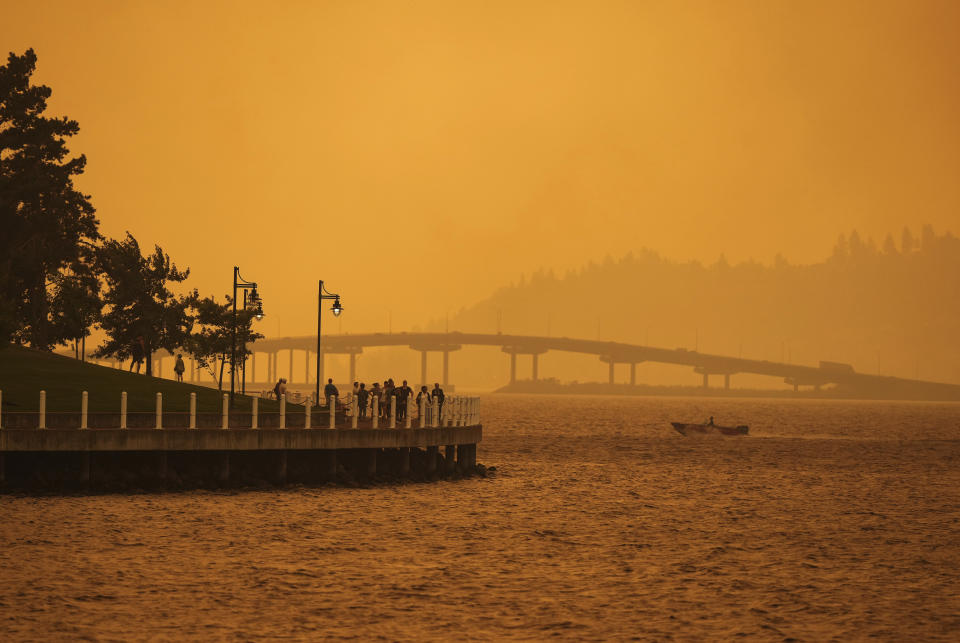 A person travels in a boat past people walking on the boardwalk as smoke from the McDougall Creek wildfire blankets the area on Okanagan Lake in Kelowna, British Columbia, Friday, Aug. 18, 2023. (Darryl Dyck/The Canadian Press via AP)