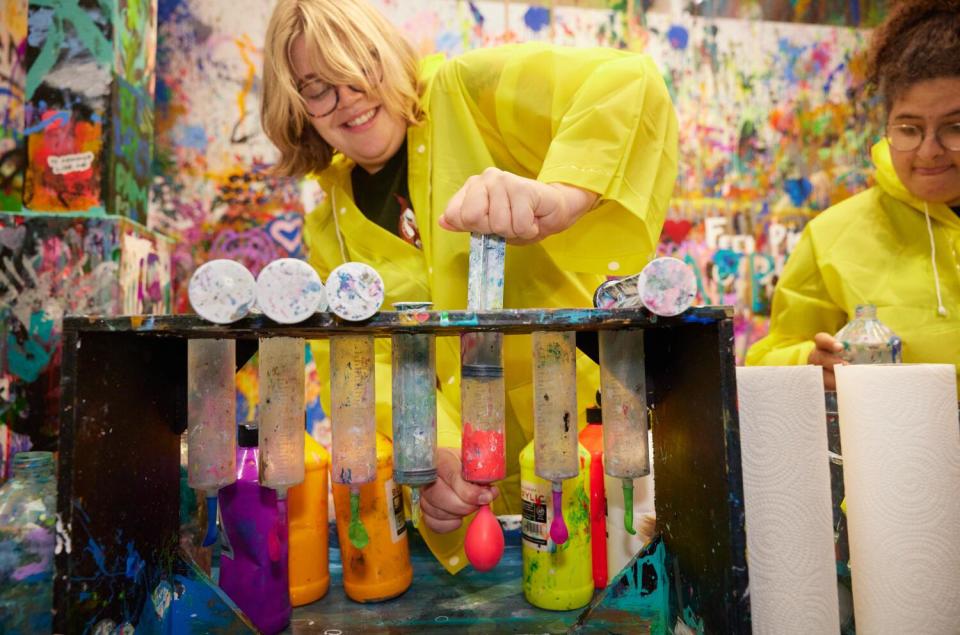 A person wearing a yellow raincoat smiles down at tubes of paint.