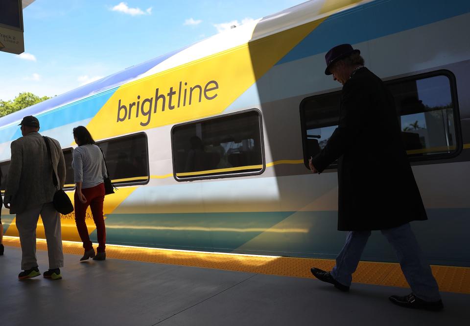 A multicolored Brightline train sits on a track with several passengers walking on the platform in front of it.