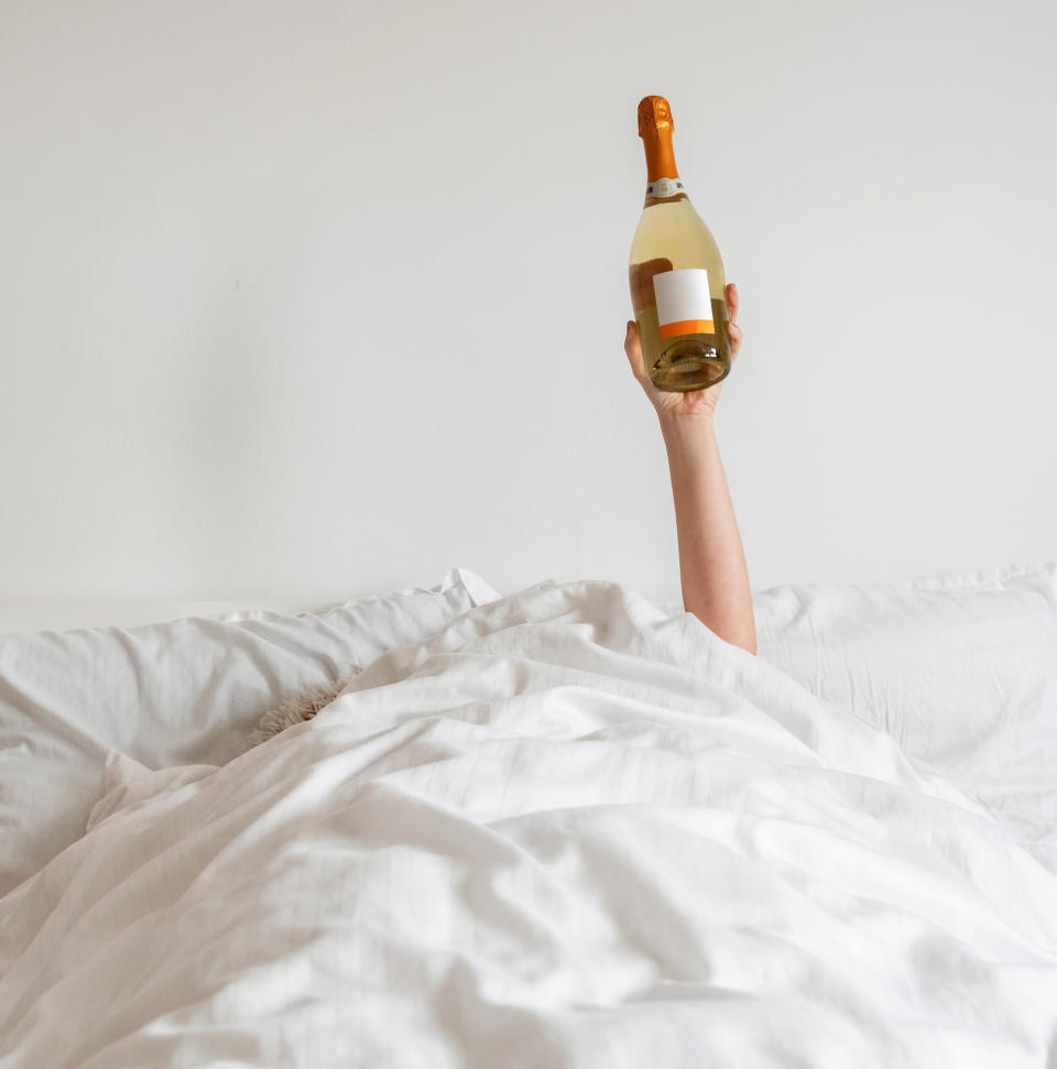 A hand holding up a bottle of wine from under the covers in a bed