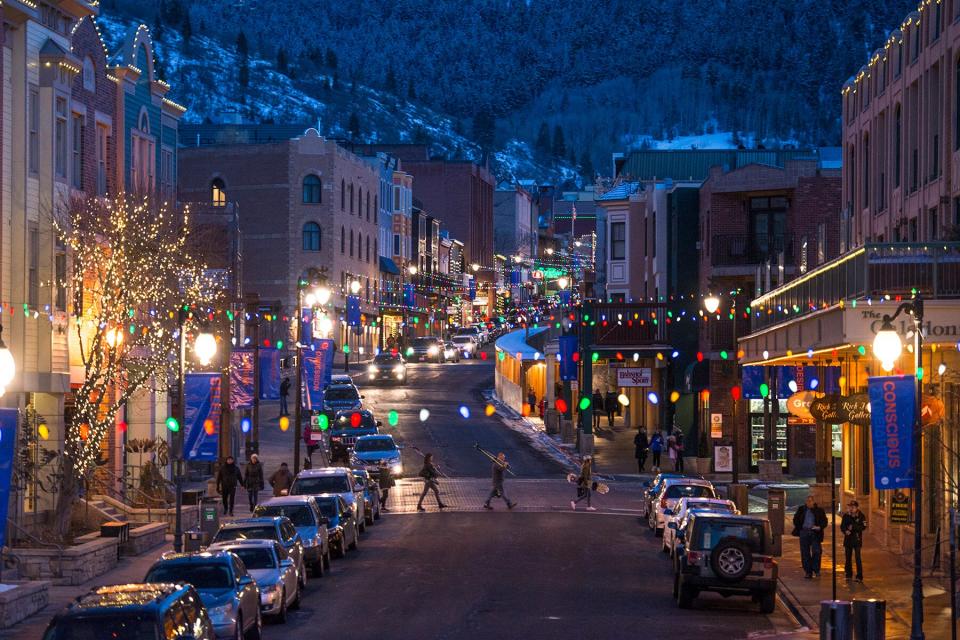 A family of ski and snowboard enthusiasts cross a festively lit and decorated Main Street Park City.