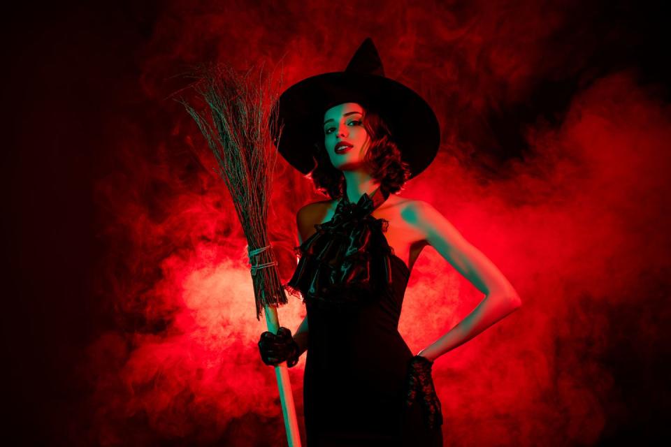 devil cruel mysterious woman wear gothic witch dress headwear holding flying broom stick isolated red fog color background