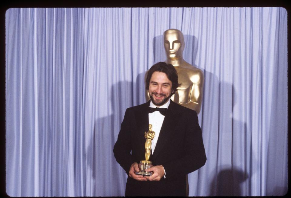 robert deniro, wearing a black tuxedo, smiling and holding an oscar statuette and standing in front of a purple curtain and a large oscar statue