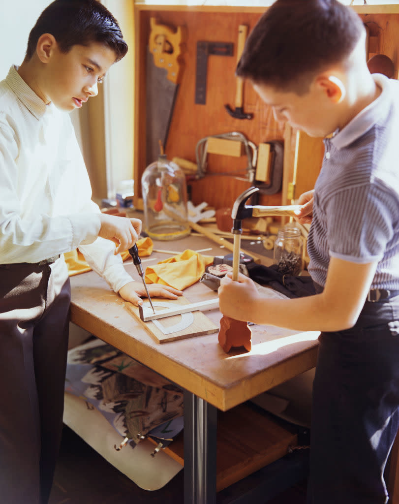 Two boys working on a woodworking project in a workshop, one using a screwdriver and the other a hammer. Various tools and materials are visible on the workbench