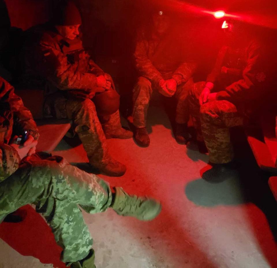 Men in military clothing sit in a dark room lit by a single red light.