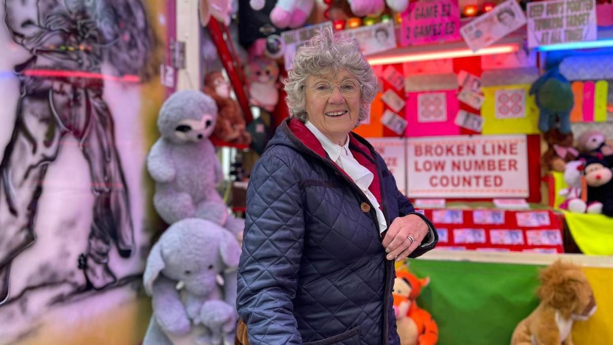 An older lady smiles in front of her rifle range. Cuddy toys, including an elephant and a tiger, are visible.