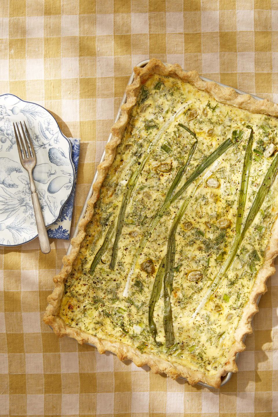 quiche made in a sheet pan set on a yellow and white checked tablecloth with a set of plates to the side