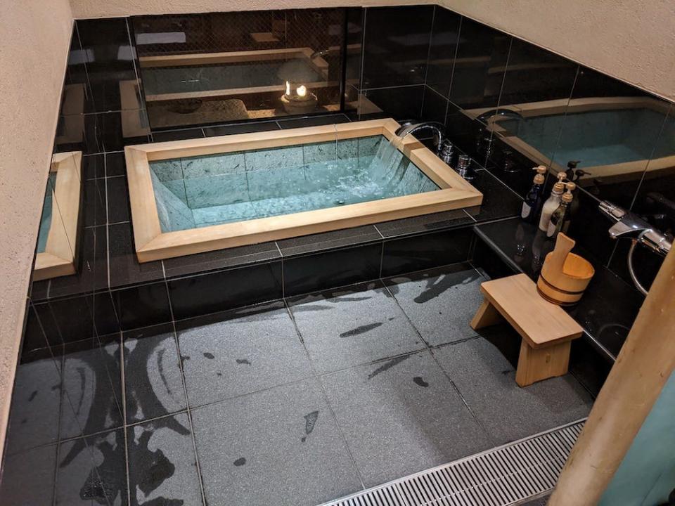 A room with black stone tiles on the floor and walls. On the left side of the room is a deep bathtub with turquoise tiles and a wooden edge. There is water coming from the tap. To the right is a small wooden stool with a wooden bucket on top, a metal shower tap, and three bottles. There is a window to the left of the bathtub. Outside is a small stone lamp with light coming out.