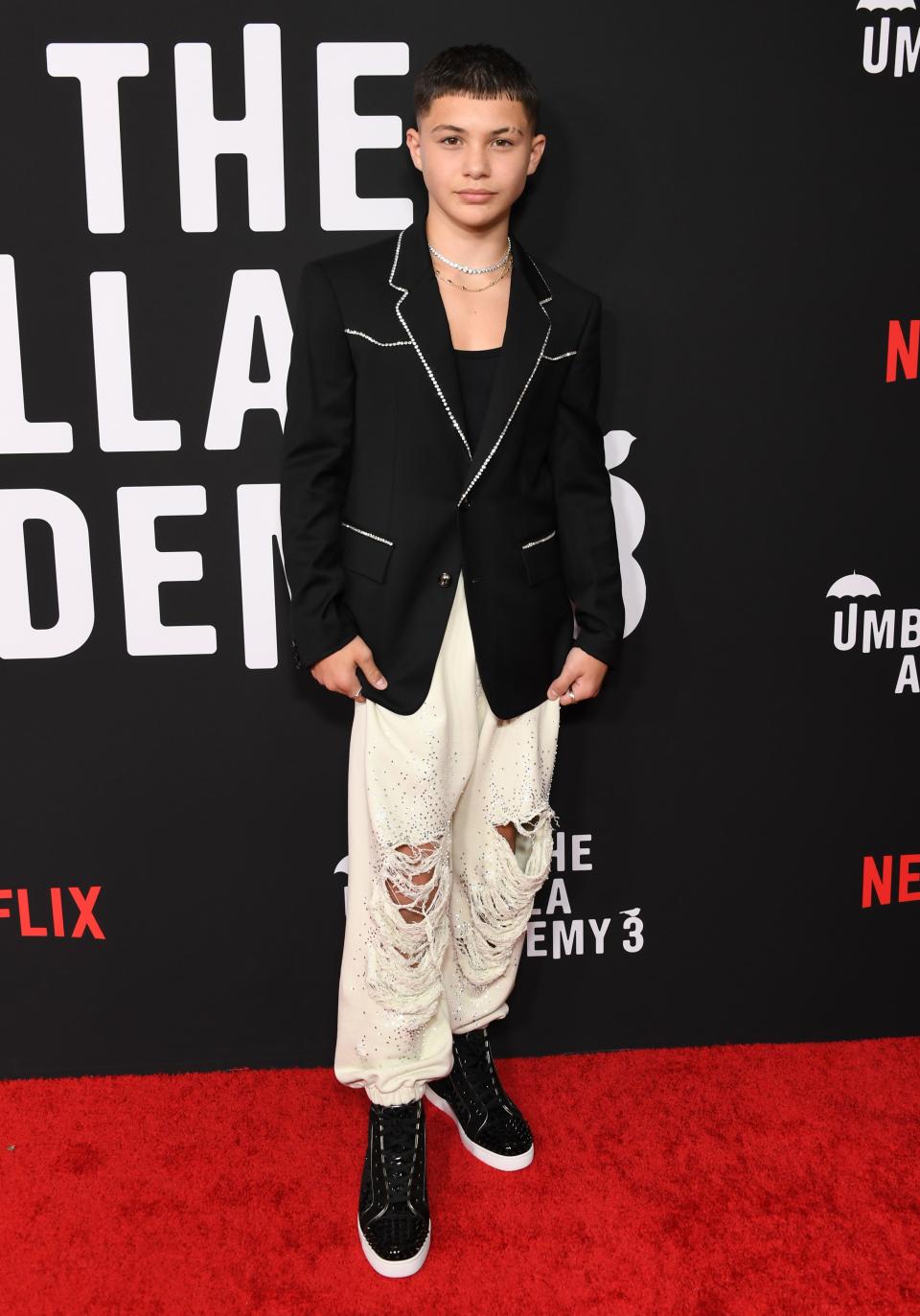 Javon in a black blazer with silver crystals along stitch lines, the collar, and pockets. He's wearing cream jogger pants with rips and crystals along with black sneakers.