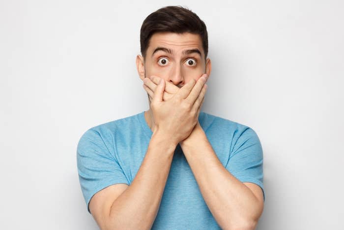 Man with shocked expression covering mouth with hands, standing against a plain background