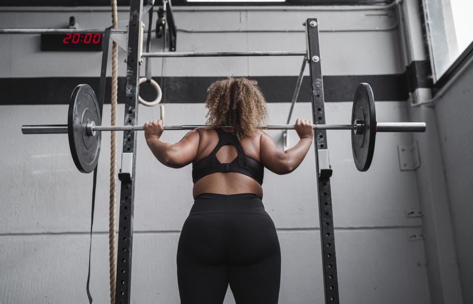 Woman performing a back squat in a gym, facing a squat rack, digital clock reads 20:00