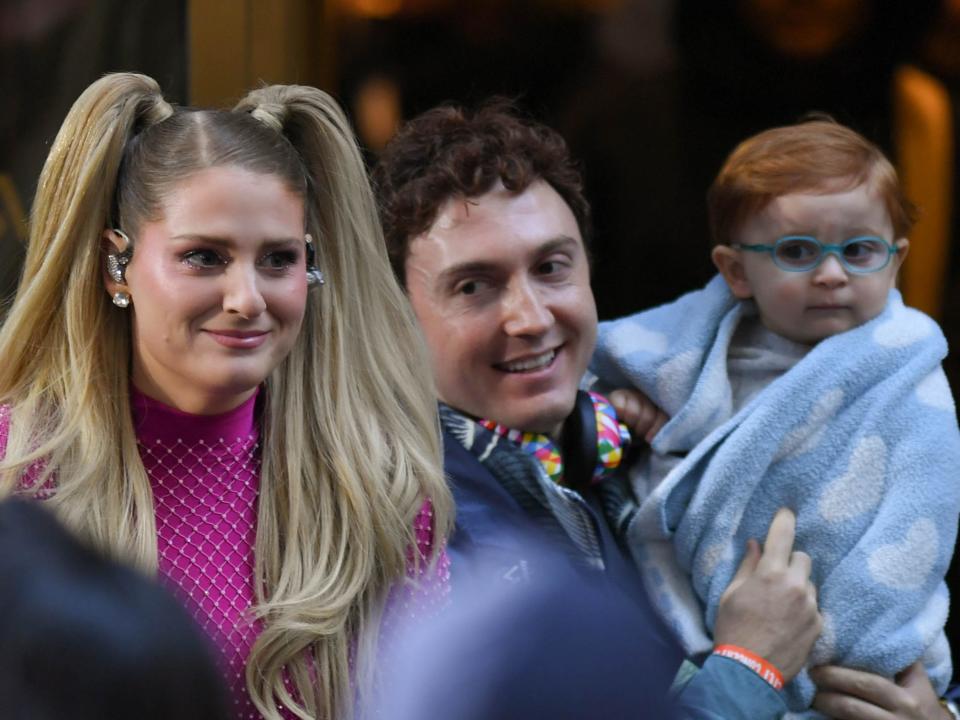 meghan trainor, in a bright pink jumpsuit and pigtails, standing next to husband daryl sabara, who is holding their baby son riley, a red-haired chiled with bright blue glasses wrapped in a light blue blanket