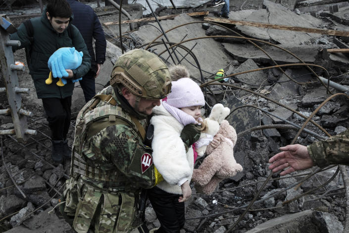 A soldier carries a child, clutching stuffed animals, through rubble.