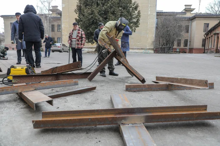 Eight people stand outside near partially completed Czech hedgehogs, as a welder holds together sections of steel beams.