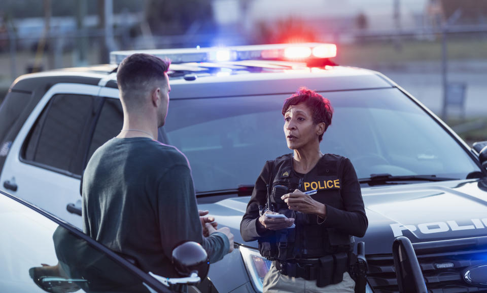 A police officer speaking to a man beside a patrol car with flashing lights