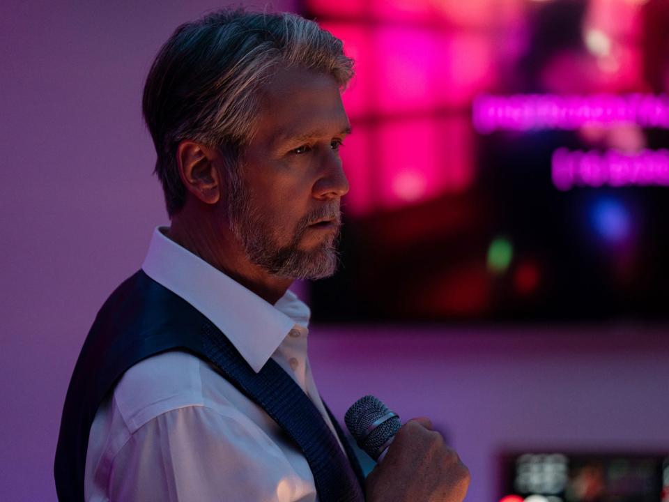 An older man wearing a suit holds a microphone sadly in a karaoke room.