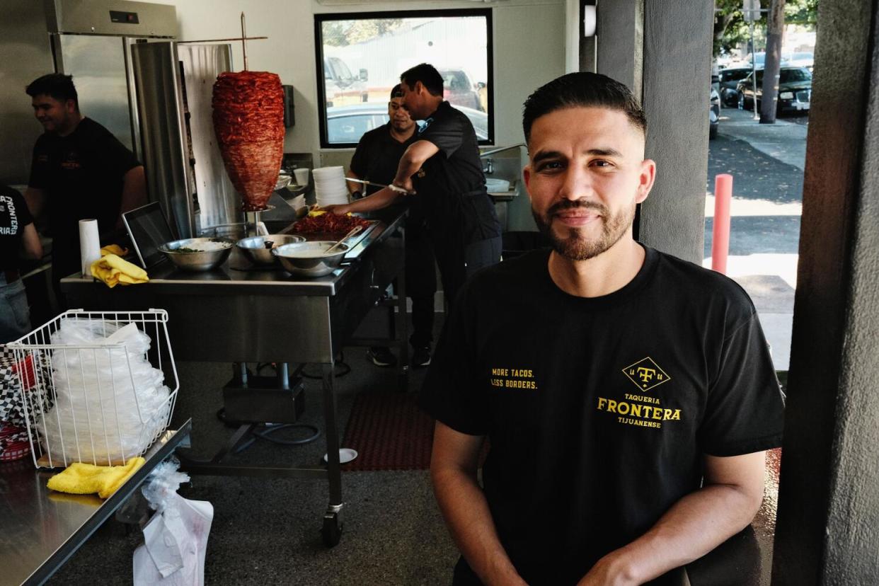 Juan Carlos "J.C." Guerra leans against an interior wall of the Taqueria Frontera kitchen. Behind, employees work.